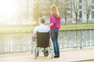 Rear View Of Woman With Her Disabled Husband On Wheelchair Looking At Lake